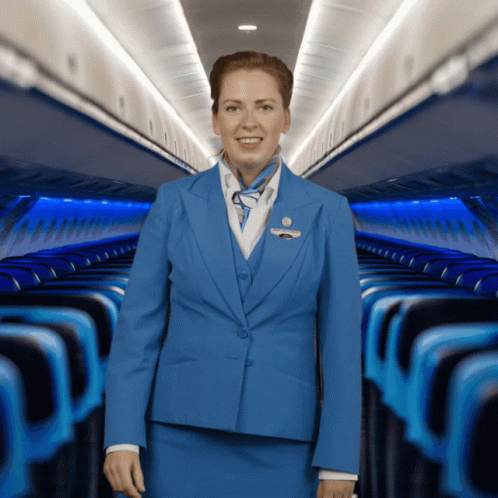 A Stewardess In A Blue Suit Is Smiling In Front Of Rows Of Blue Seats GIF
