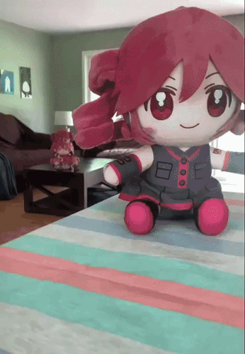 A Stuffed Doll With Red Hair Is Sitting On A Striped Blanket GIF