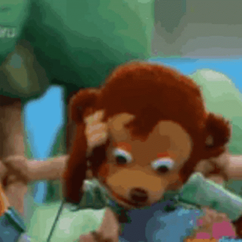 A Stuffed Monkey Is Scratching His Head While Sitting On A Green Blanket . GIF