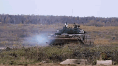A Tank With The Number 3 On The Side Is Driving Through A Grassy Field . GIF