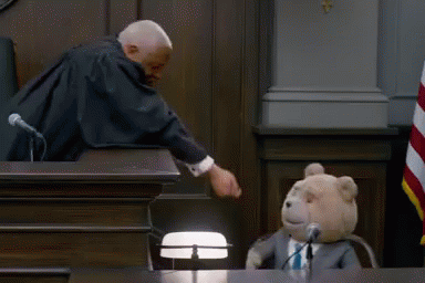 A Teddy Bear In A Suit And Tie Is Sitting In Front Of A Judge In A Courtroom GIF