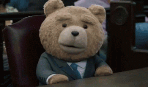 A Teddy Bear In A Suit And Tie Sits At A Table GIF