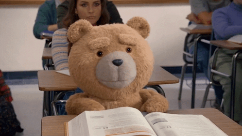 A Teddy Bear Sits At A Desk In A Classroom Reading A Book GIF