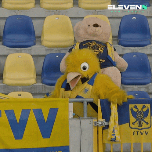 A Teddy Bear Sits Next To A Mascot In A Stadium GIF