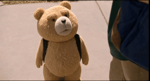 A Teddy Bear Wearing A Backpack Stands On A Sidewalk GIF