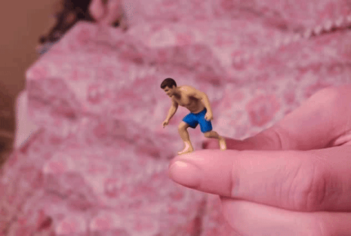 A Tiny Figure Of A Man In Blue Shorts Is Being Held By A Pink Finger GIF
