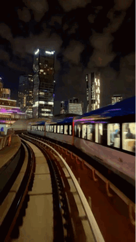 A Train Is Going Down The Tracks At Night With Buildings In The Background GIF