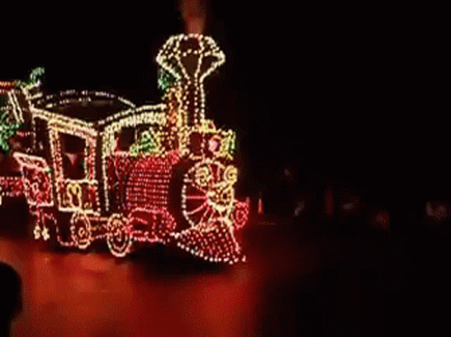 A Train With Christmas Lights On It Is Driving Down A Street GIF