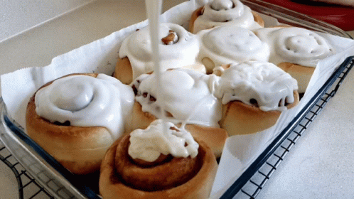 A Tray Of Cinnamon Rolls With White Frosting Being Poured On Them GIF