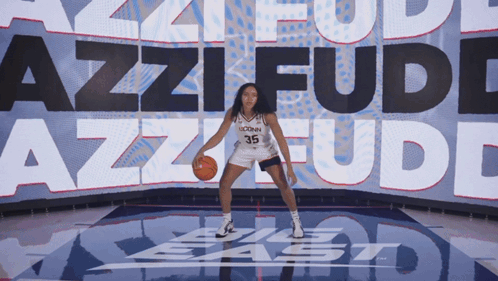 A Uconn Basketball Player Dribbles A Ball In Front Of A Large Screen That Says Azzi Fudd GIF