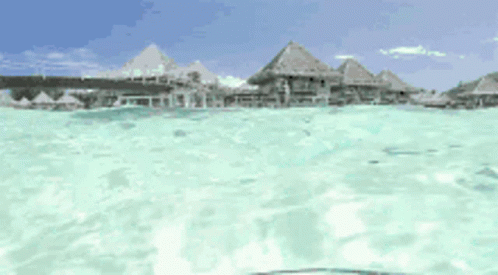 A View Of A Tropical Island From The Water With Thatched Huts In The Background GIF