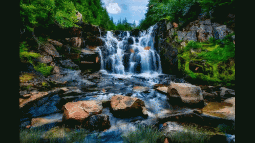 A Waterfall Surrounded By Rocks And Trees With A Person Standing In The Background GIF