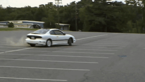 A White Car Is Drifting In A Parking Lot With A Building In The Background GIF