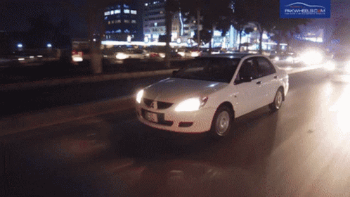 A White Car Is Driving Down A Street At Night With A Pakwheels.com Logo Behind It GIF