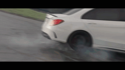 A White Car Is Driving Down A Wet Road With Smoke Coming Out Of The Tire GIF