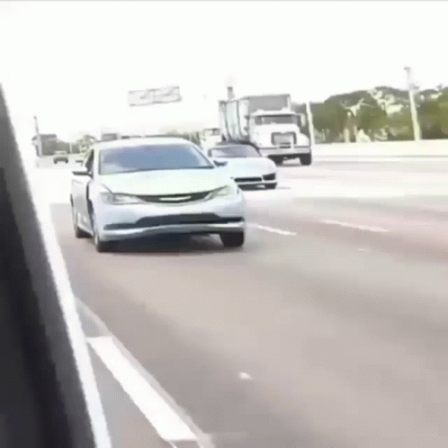 A White Car Is Driving Down The Highway Next To A Truck . GIF