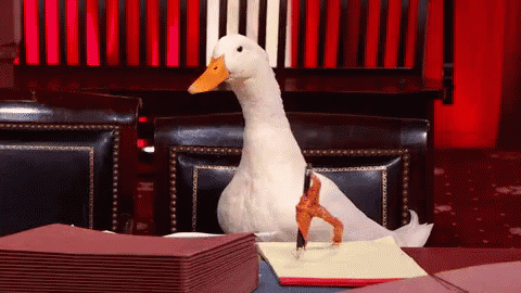 A White Duck Is Sitting At A Table With A Pen In Its Foot GIF