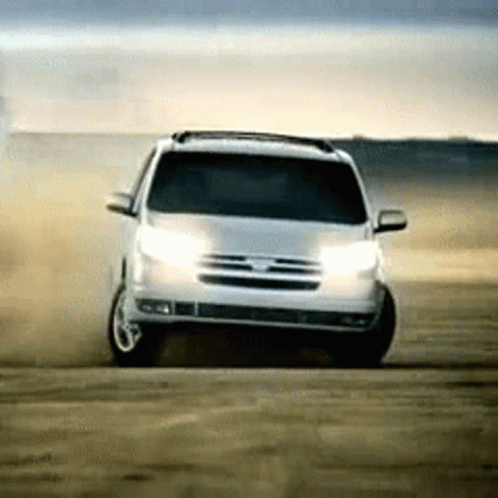 A White Suv Is Driving On A Dirt Road With Its Lights On GIF