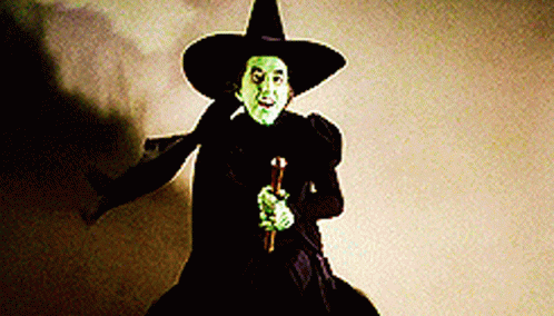 A Witch With Green Eyes And A Black Hat Is Holding A Cane . GIF