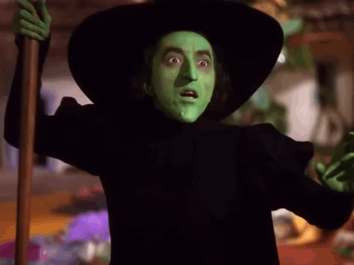 A Witch With Green Paint On Her Face Is Holding A Cane GIF