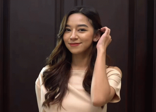 A Woman Adjusts Her Hair In Front Of A Dark Wooden Wall GIF