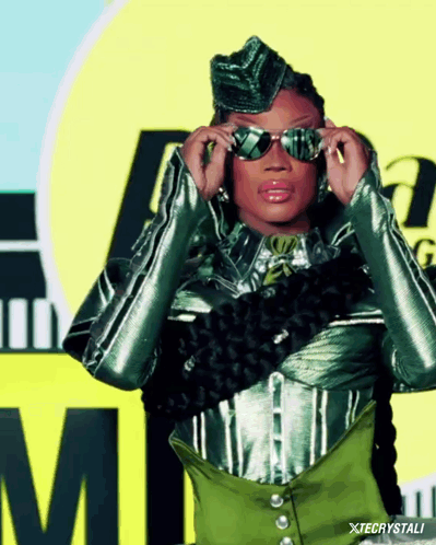 A Woman Adjusts Her Sunglasses In Front Of A Yellow Sign With The Letter M On It GIF