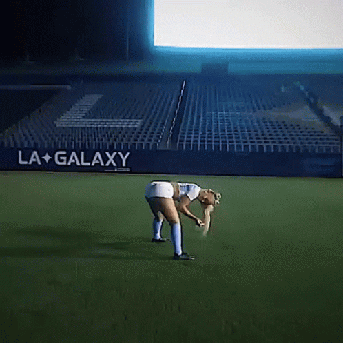 A Woman Bending Over On A Soccer Field In Front Of A La Galaxy Banner GIF