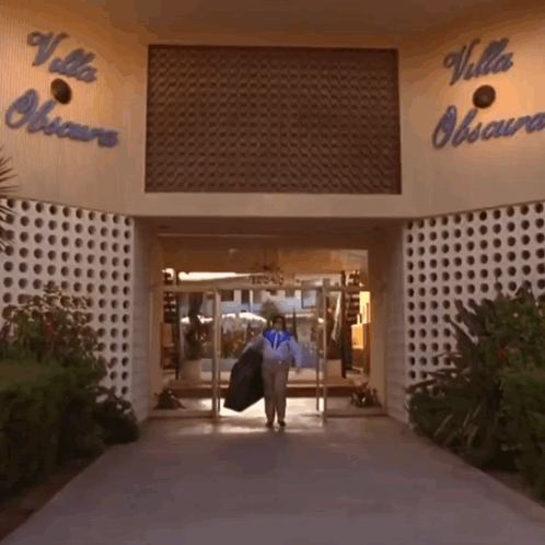 A Woman Carrying A Jacket Walks Into The Villa Obscura Building GIF