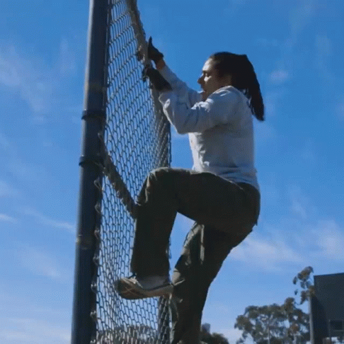 A Woman Climbs A Chain Link Fence Against A Blue Sky GIF