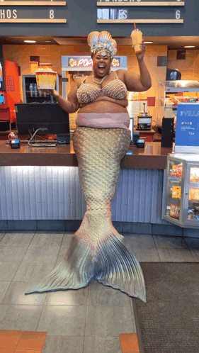 A Woman Dressed As A Mermaid Holds A Drink In Front Of A Pop Corn Machine GIF