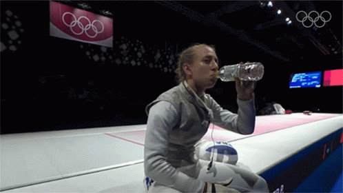 A Woman Drinking Water From A Bottle In Front Of A Sign That Says Olympic Rings GIF