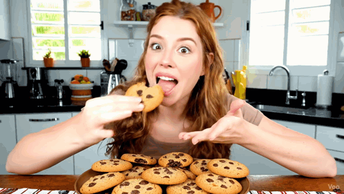 A Woman Eating A Chocolate Chip Cookie From A Plate Of Cookies GIF