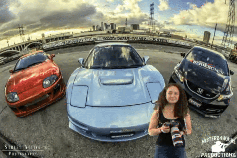 A Woman Holding A Camera Stands In Front Of A Series Only Car And A Red Car GIF