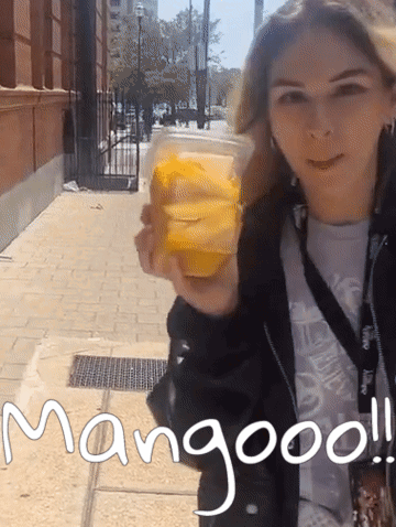A Woman Holding A Cup Of Mango Juice With The Words Mangooo Written On The Sidewalk GIF