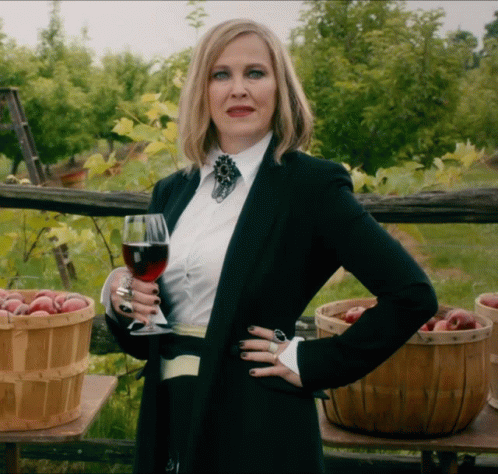 A Woman Holding A Glass Of Wine In Front Of Baskets Of Fruit GIF