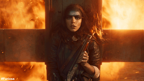 A Woman Holding A Gun In Front Of A Cross With The Hashtag #Furiosa GIF