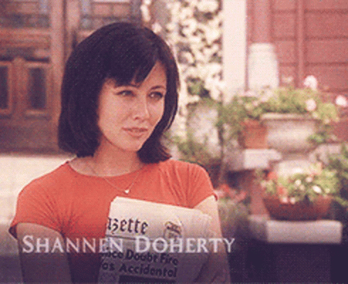 A Woman Holding A Newspaper With The Name Shannon Doherty Written On It GIF
