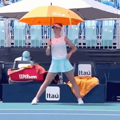 A Woman Holding An Orange Umbrella On A Tennis Court With A Wilson Bag In The Background GIF