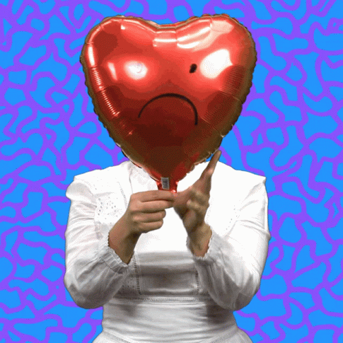 A Woman Holds A Red Heart Shaped Balloon With A Sad Face On It GIF
