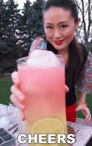 A Woman Holds Up A Glass Of Pink And Yellow Liquid With The Words Cheers Written Below Her GIF