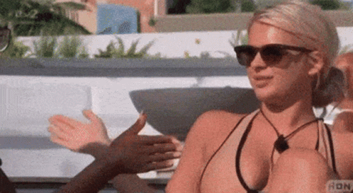 A Woman In A Bikini And Sunglasses Is Sitting On A Couch Talking To Another Woman . GIF