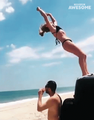 A Woman In A Bikini Is Doing A Handstand While A Man Drinks From A Cooler On The Beach GIF