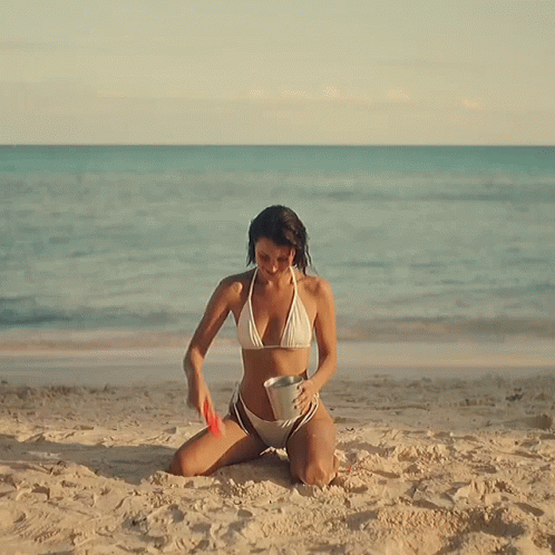 A Woman In A Bikini Is Kneeling Down On The Beach GIF