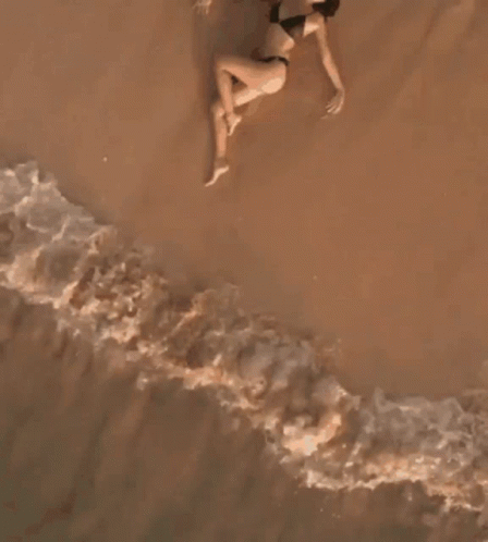 A Woman In A Bikini Is Laying On The Beach Near The Water GIF