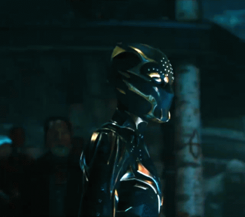 A Woman In A Black And Gold Costume With A Helmet On Stands In A Dark Room GIF