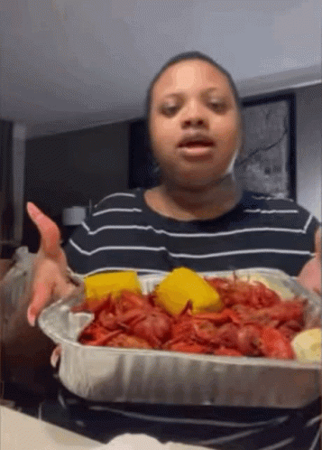 A Woman In A Black And White Striped Shirt Is Holding A Tray Of Crawfish And Cornbread GIF