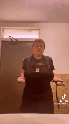A Woman In A Black Apron Is Standing In Front Of A Sink In A Bathroom . GIF