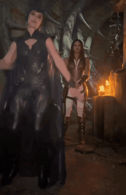 A Woman In A Black Cape Stands Next To A Woman In A White Dress GIF