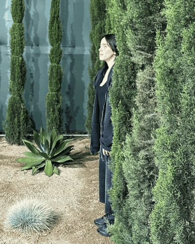 A Woman In A Black Coat Is Standing Behind A Bush GIF