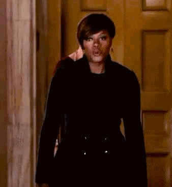 A Woman In A Black Coat Is Standing In Front Of A Door Making A Funny Face . GIF